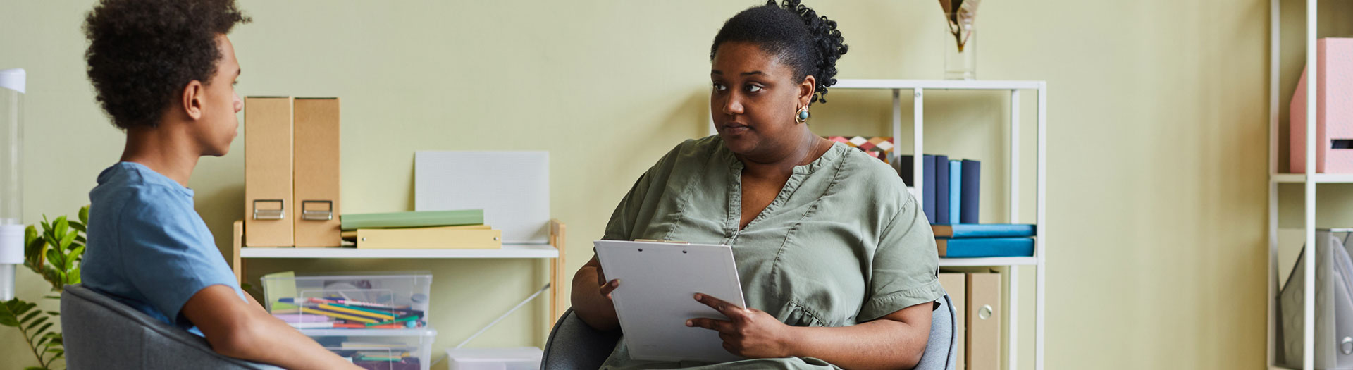 A young person sits facing to the left in a grey chair, appearing to be in conversation with an adult woman on the right, who is attentively holding a clipboard.