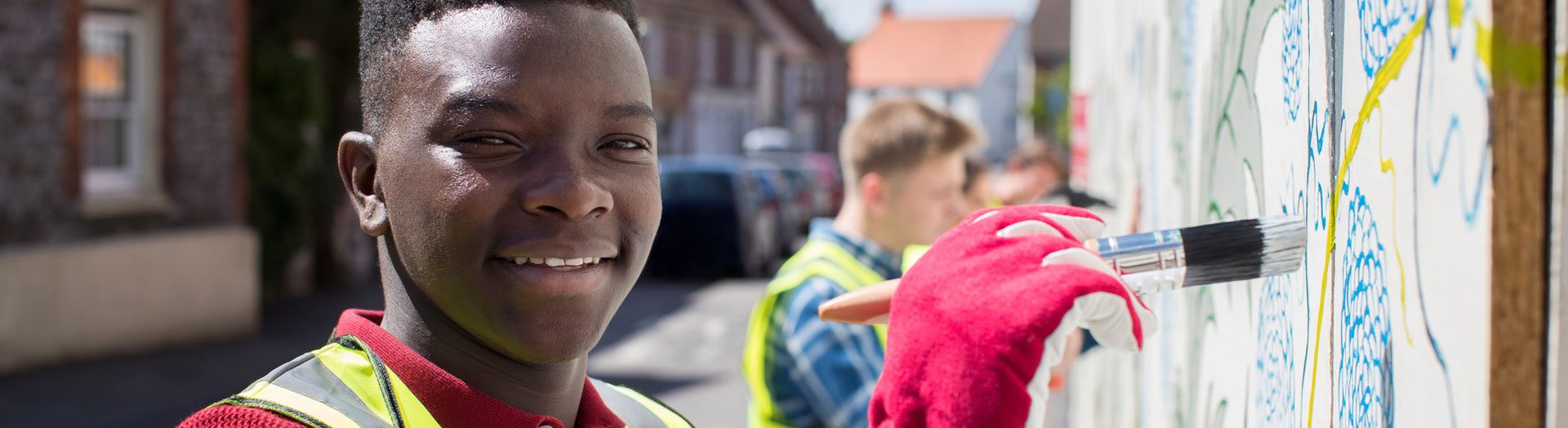 A smiling young person in a reflective safety vest holds a paintbrush up to a mural, adding vibrant colors to the artwork. In the background, another person in a similar vest is focused on painting, while a residential street scene fills the sunny background.