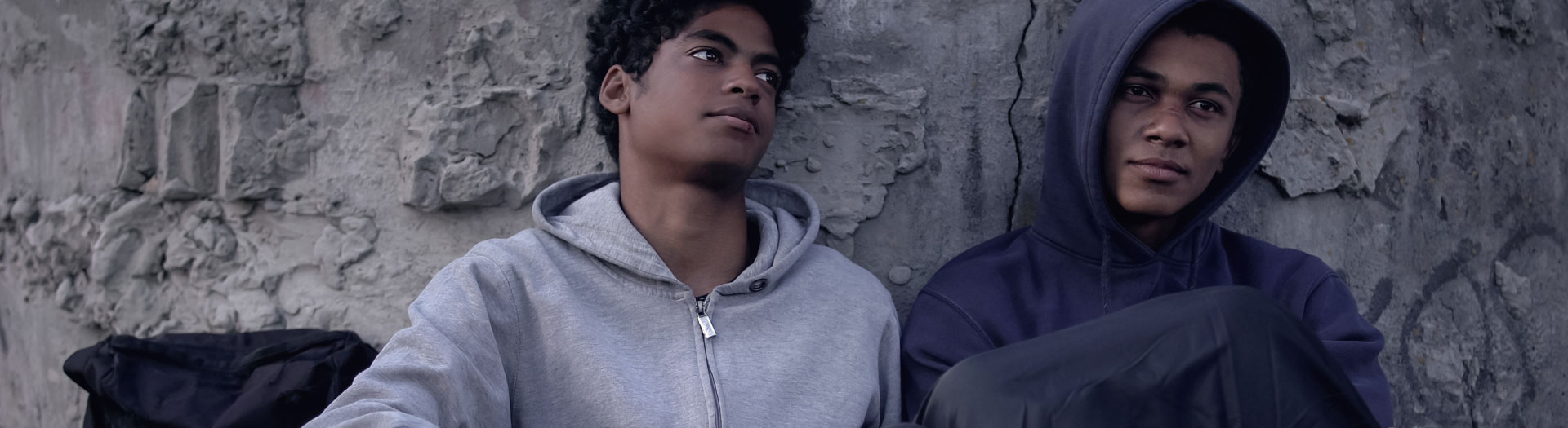 Two young men leaning against a textured concrete wall, one wearing a light gray hoodie and the other in a dark blue hoodie. Both are gazing off into the distance with thoughtful expressions.