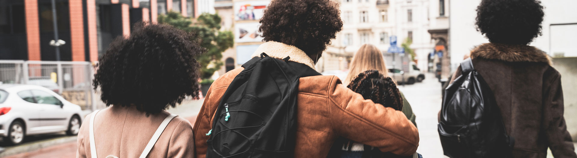 "Four people from behind walking on a city street, focused on the first three individuals with curly hair and diverse styles, one wearing a tank top, another in a brown leather jacket, and the third in a fur-lined coat.