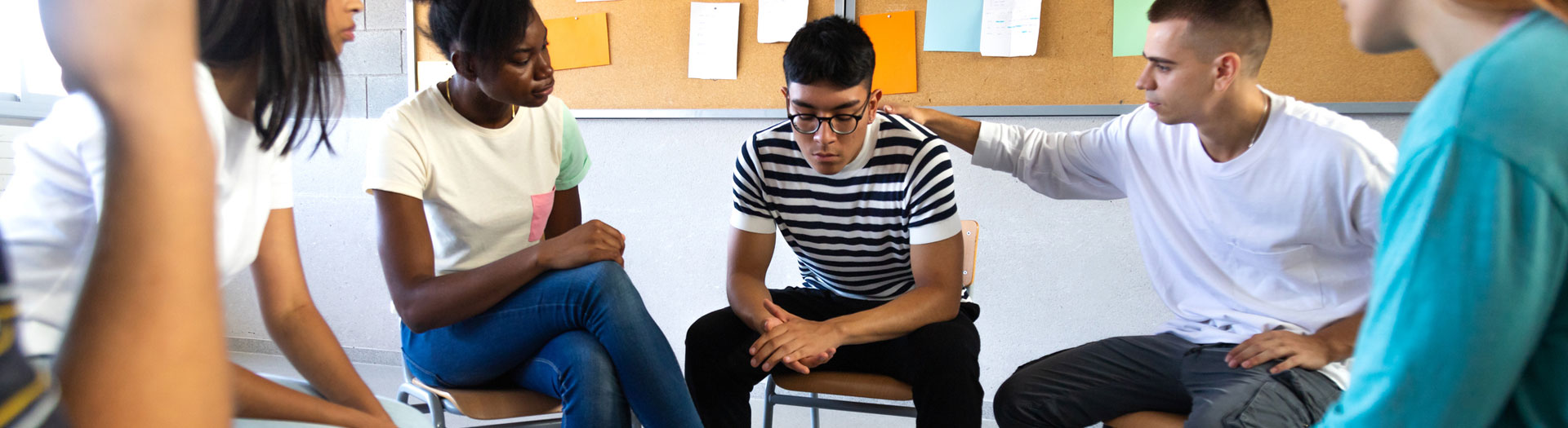 A diverse group of young adults seated in a circle, engaging in what appears to be a supportive discussion. One person in a striped shirt seems to be the focus of attention, looking down thoughtfully while another participant is placing a comforting hand on their shoulder.