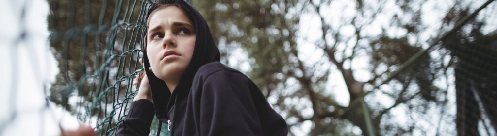 A pensive young girl with a hooded sweatshirt is leaning against a chain-link fence, gazing upward with a contemplative expression.