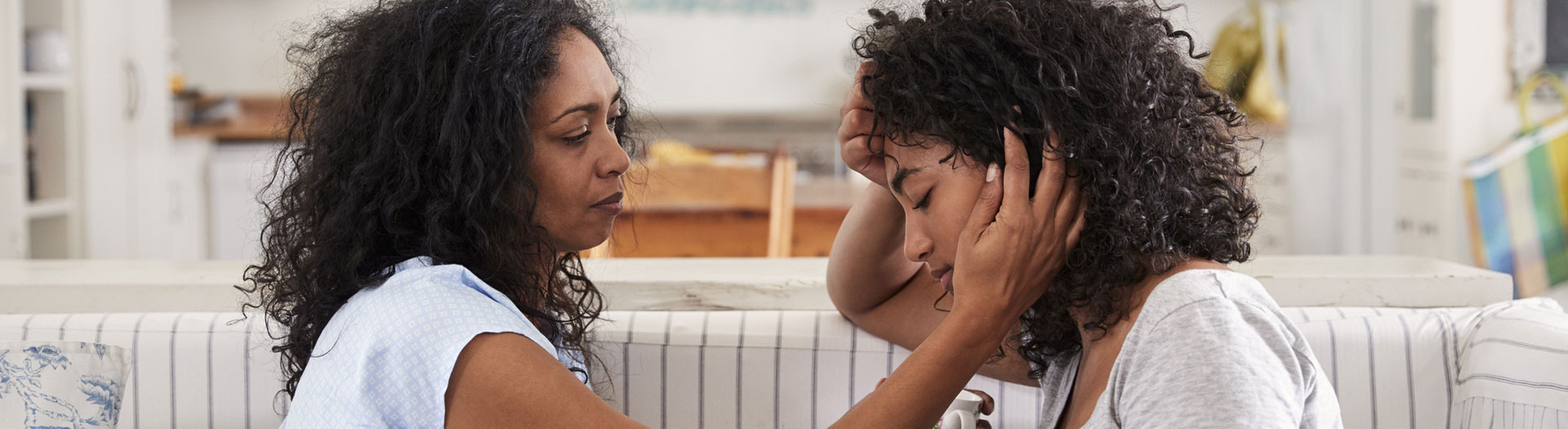 A compassionate scene of two women in a comforting embrace. The older woman, wearing a blue top, is holding the younger woman's head tenderly, who is in a grey top with her eyes closed, showing signs of distress or sadness.
