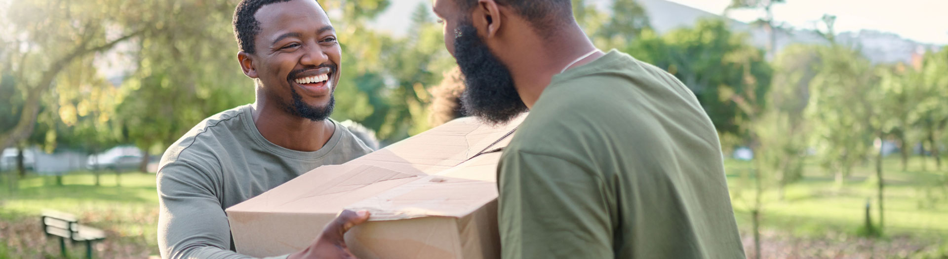 Two happy men exchanging a large cardboard box outdoors in a sunny park.