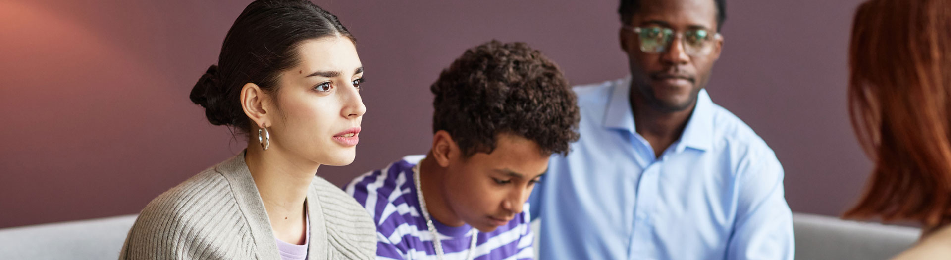 A focused woman with her hair in a bun, wearing a cardigan, listens intently in a meeting alongside a boy in a striped shirt who is looking down, and a man in a blue shirt with glasses