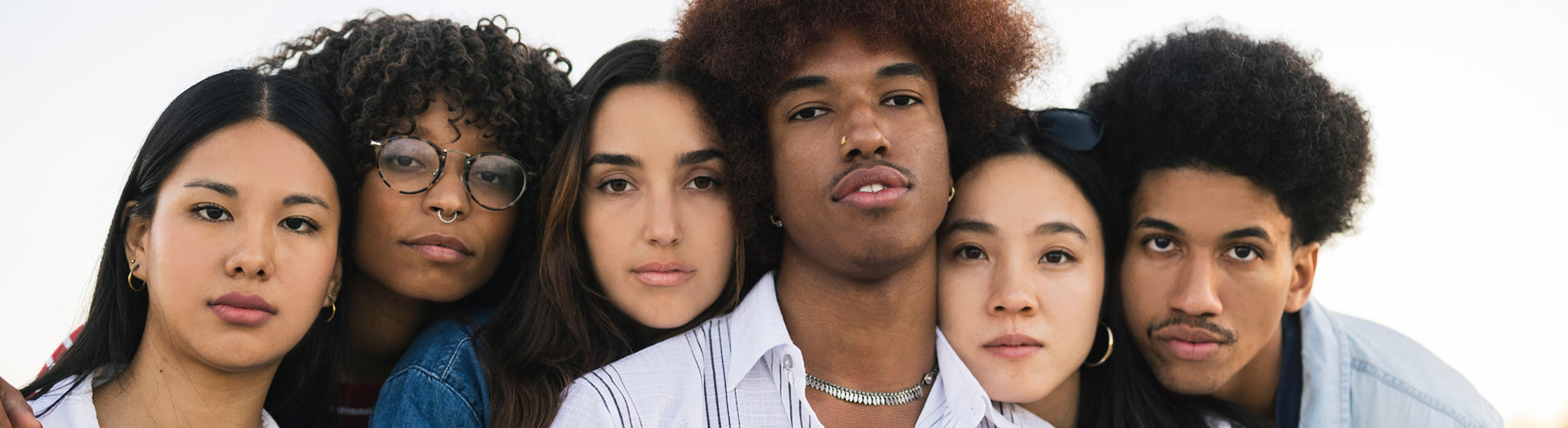 A close-up of a diverse group of five young people with different hairstyles and features, standing shoulder to shoulder and facing the camera against a clear sky.