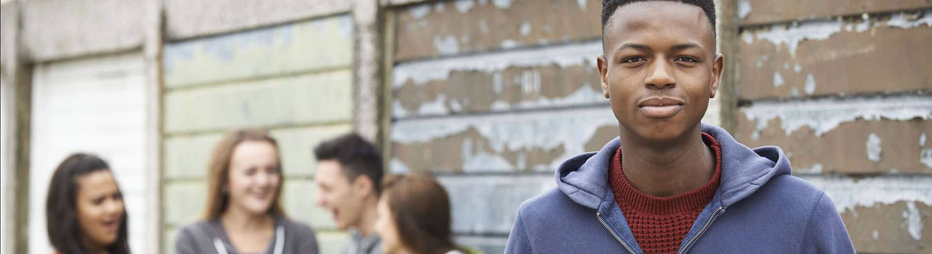 A confident young man in the foreground looks directly at the camera, with a slight smile, wearing a blue hoodie and a maroon sweater underneath. In the blurred background, a group of people is laughing and interacting with each other.