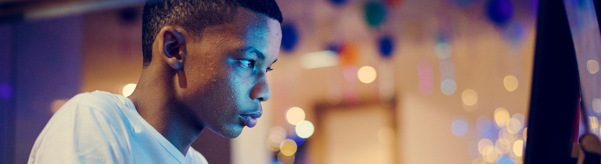 A young man in profile, wearing a white t-shirt, looks intently at something off-camera. The background is filled with soft, out-of-focus lights, suggesting an indoor or nighttime setting.