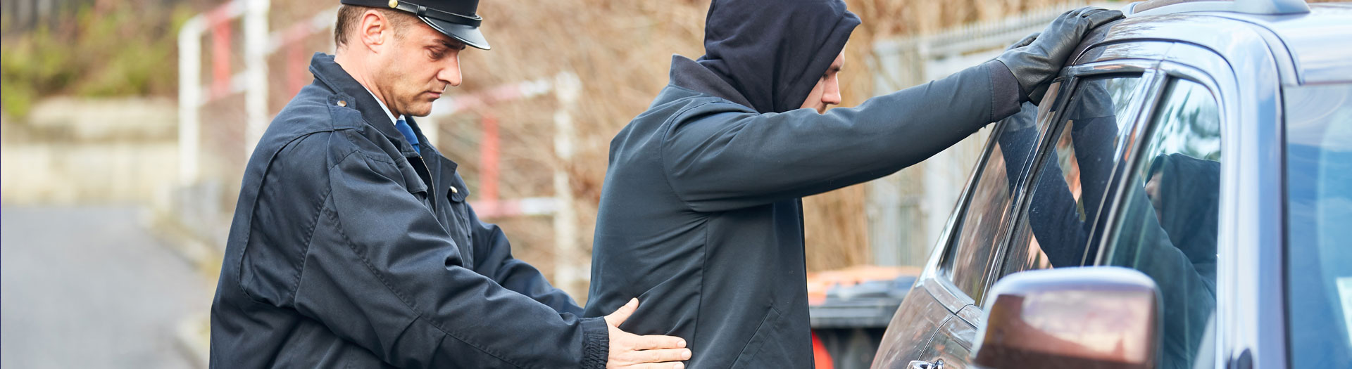 A police officer is conducting a pat-down on a young man in a black hoodie who is leaning forward onto a car, suggesting a stop-and-search procedure in an urban outdoor setting.