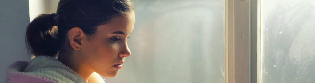 A young woman with brown hair tied back, wearing a white bathrobe, looks thoughtfully out of a window with a soft, contemplative expression. Natural light illuminates her face.