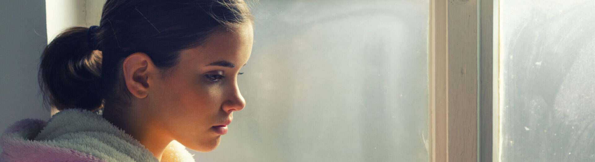 A young woman with brown hair tied back, wearing a white bathrobe, looks thoughtfully out of a window with a soft, contemplative expression. Natural light illuminates her face.